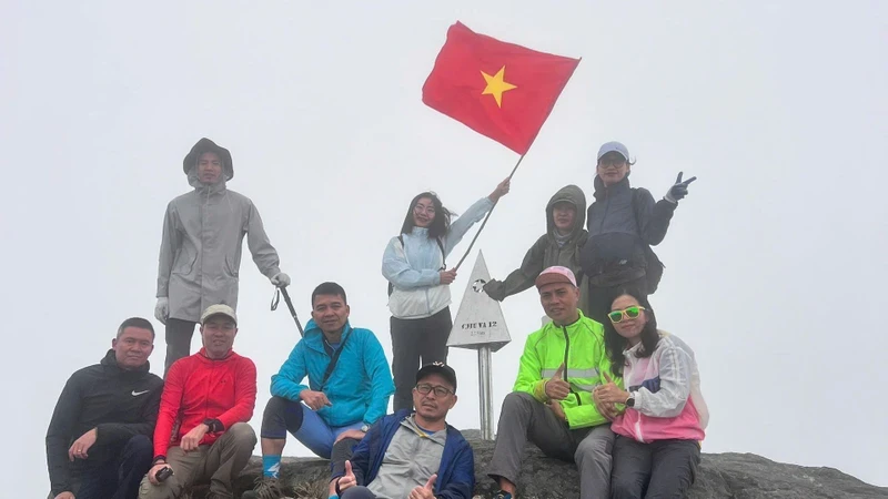 A group of tourists pose with the metal pyramid marked “Chu Va 2,751m” amidst howling winds.